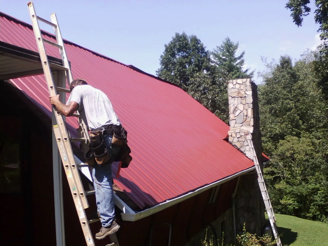 Roofer on a ladder beside a red metal roof and stone chimney — Soffit & Fascia Repair by Roofing Brothers in Atkins